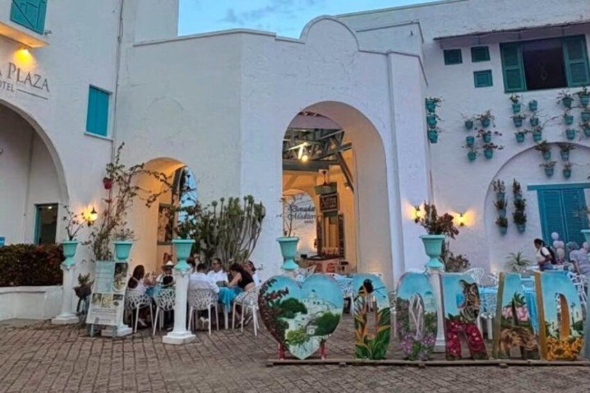 A white building with arches and a sign that reads 'Plaza Hotel.' There are potted plants and decorative elements around the entrance. 4