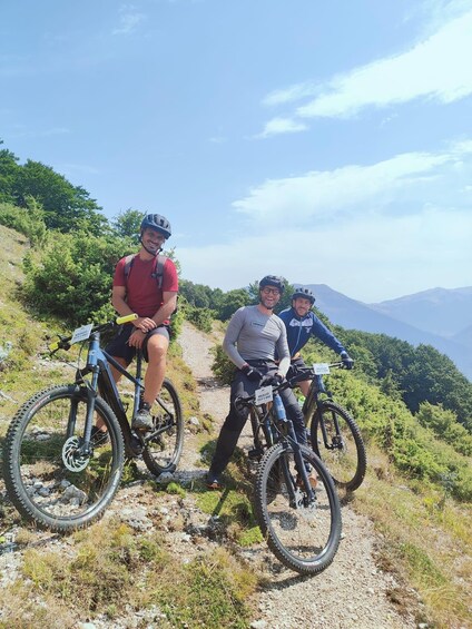 Three cyclists on a mountain trail with a clear sky and distant mountains. 1