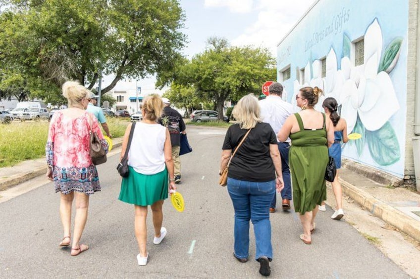 A group of people walking along a street with a mural of white flowers on the wall. 4