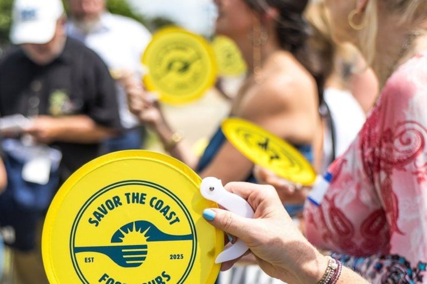 A person holding a yellow frisbee with the text 'SAVOR THE COAST' and 'EST 2025' on it. 2