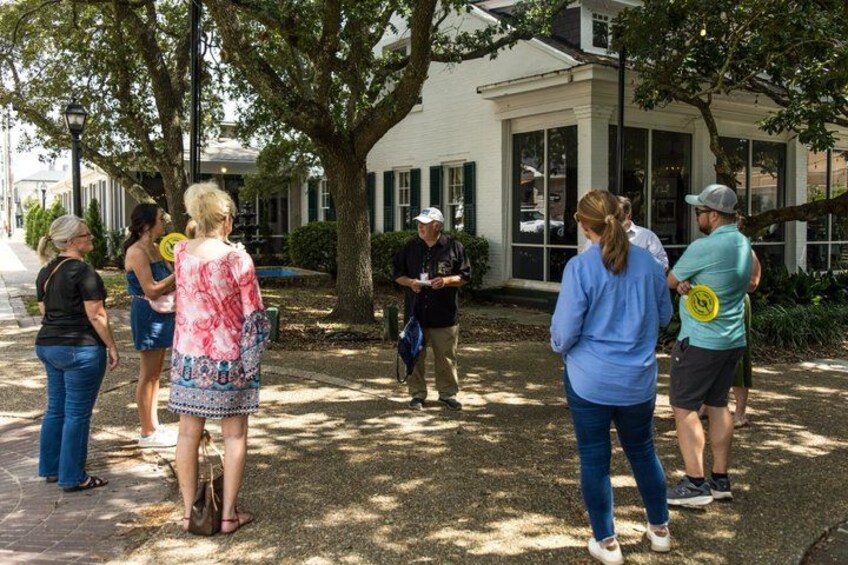 A group of people standing outdoors, some holding frisbees, in a shaded area with trees and a building in the background. 5