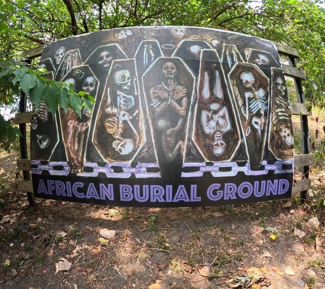 Un letrero con el texto 'African Burial Ground' rodeado de árboles. 3