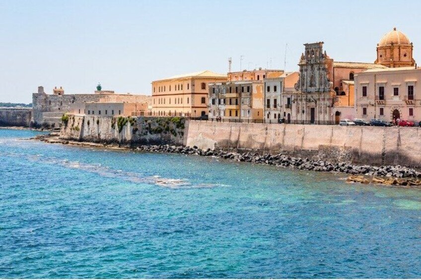 A coastal cityscape with historic buildings, a clear blue sea, and a fortified wall. 4