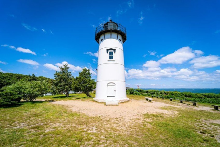 A white lighthouse with a black top, surrounded by a grassy area with trees and a clear blue sky. 1
