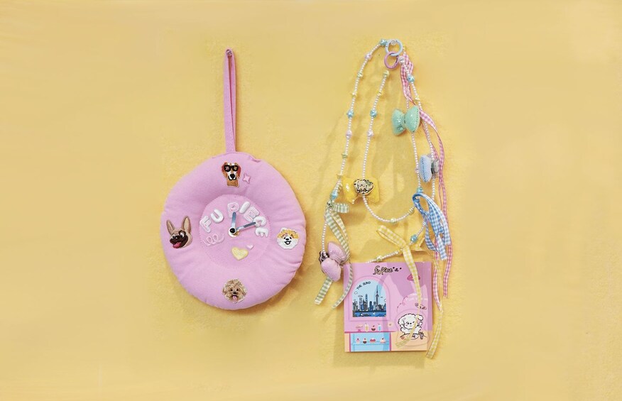 A pink plush toy with a clock face and various animal faces, a pink and blue striped keychain, and a small card with a cityscape illustration. 4