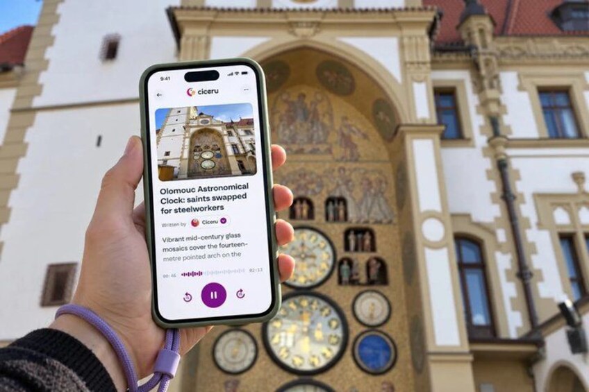 A smartphone displaying an article about the Olomouc Astronomical Clock, with a historic building in the background. 1