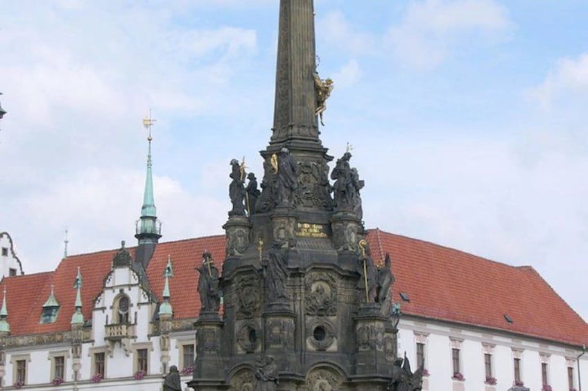 A historic monument with intricate carvings and statues, set against a backdrop of traditional architecture with red-tiled roofs and a church with a green spire. 4