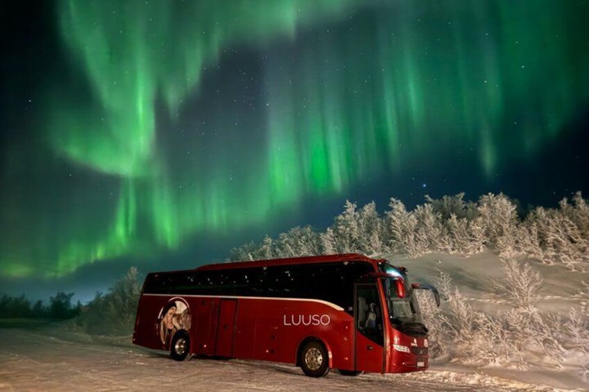 A red bus with 'LUUSO' on the side parked on a snowy road with the aurora borealis in the background. 1