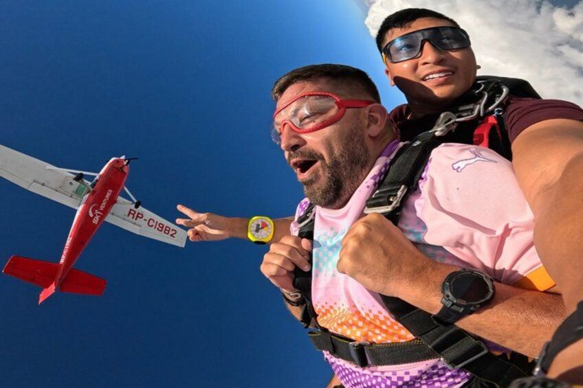 A person in a pink shirt and a person in a purple shirt with a backpack, both looking at a red airplane with the registration RP-C18B2 in the sky. 4