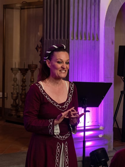 Une personne vêtue d’une robe rouge ornée de broderies blanches, se tenant dans une pièce éclairée d’une lumière violette, avec un pied de micro et une grande fenêtre dotée de stores verticaux. 3