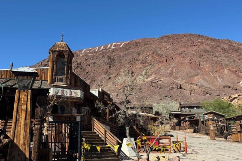 A rustic wooden building with a bell tower, a sign reading 'R D,' and a mountainous backdrop. 1