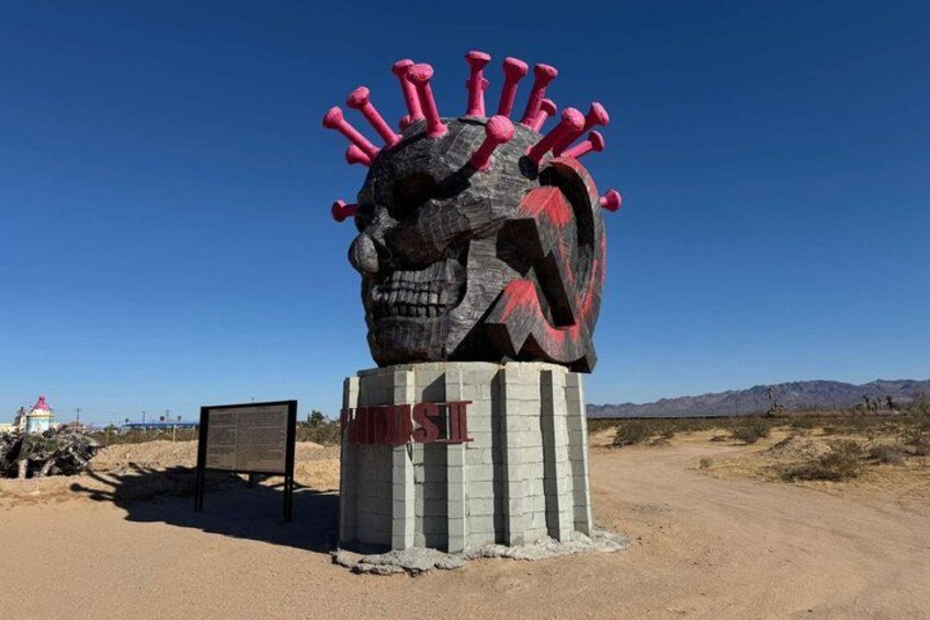 A large skull sculpture with a red crown, located on a dirt road with a clear blue sky and distant mountains. 3