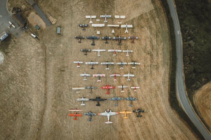 An aerial view of a large collection of aircraft parked in rows on a dry, grassy field near a road. 1