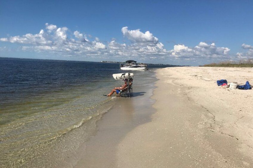 A beach with a person sitting on a chair, a boat in the water, and a sandy shore with some items on the right. 2