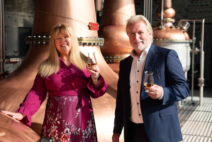 Two people holding glasses of a beverage in a distillery with copper tanks. 2