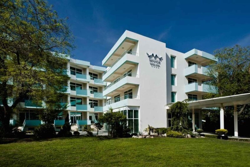 Un edificio moderno de varios pisos con fachada blanca y detalles azules, rodeado de vegetación y árboles. 1