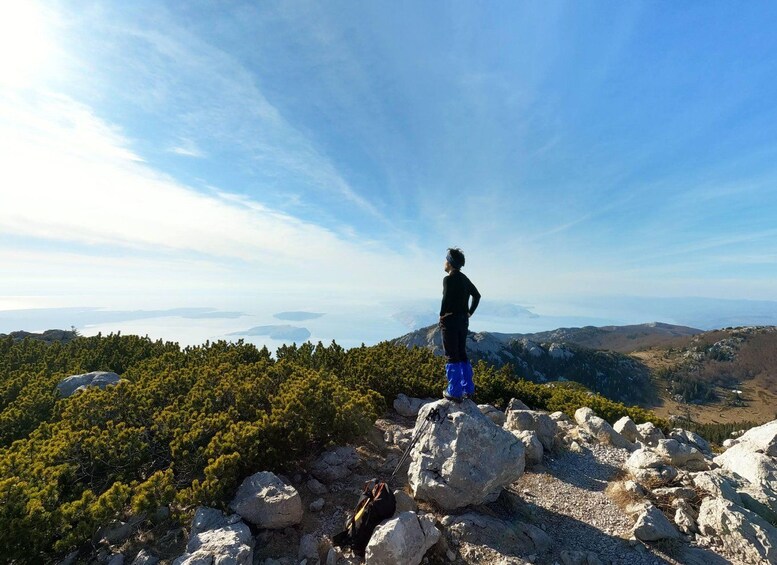 A person standing on a rocky outcrop overlooking a mountainous landscape with dense forests and a clear sky. 1
