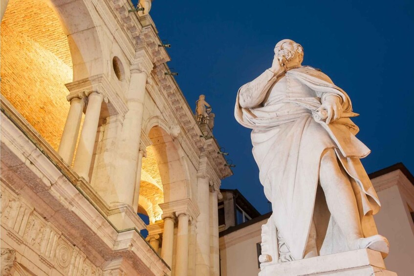 A classical statue of a seated figure with a thoughtful pose, set against a backdrop of an ornate building with arches and columns. 3