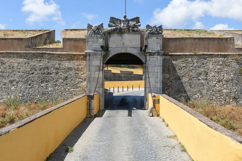 A stone archway with statues, leading through a yellow gate into a rocky area. 3