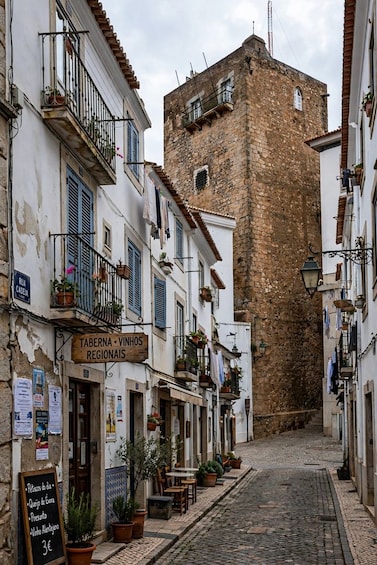 A narrow cobblestone street lined with white buildings, a stone tower, and outdoor seating areas with potted plants. 2