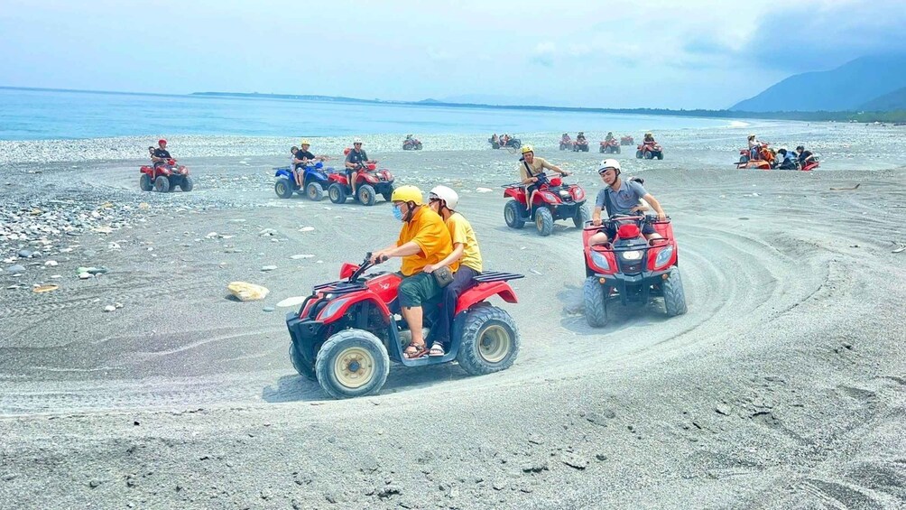 People riding ATVs on a sandy beach with a body of water and mountains in the background. 2