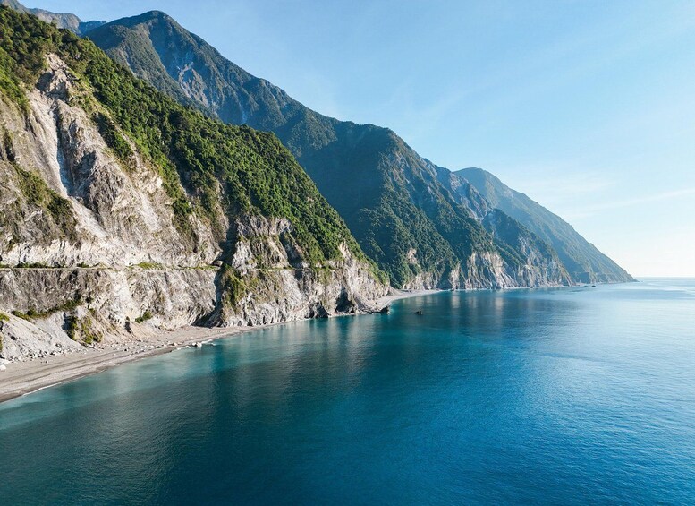 A coastal landscape with a rocky shoreline, a clear blue sea, and a mountain range in the background. 4