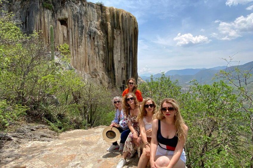 A group of people sitting on a rocky outcrop with a large cliff in the background. 1
