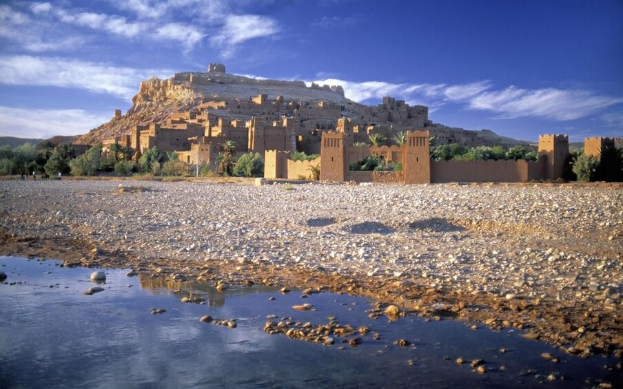 A historic fortified city with a prominent hilltop castle, a rocky riverbed, and sparse vegetation. 3