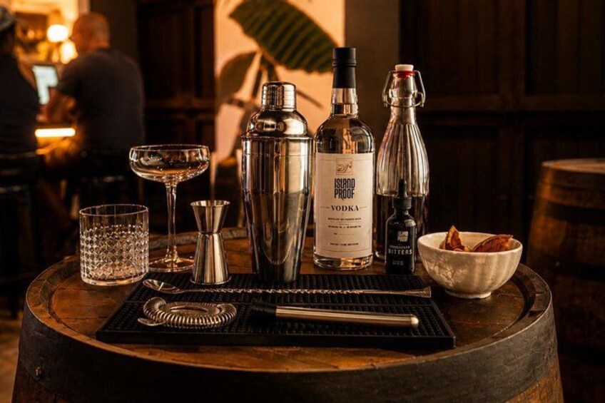 A bar setting with a bottle of vodka, a cocktail shaker, a glass, and a bowl of snacks on a wooden barrel. 1