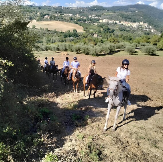 A group of people horseback riding on a dirt path through a hilly landscape with trees and buildings in the distance. 2