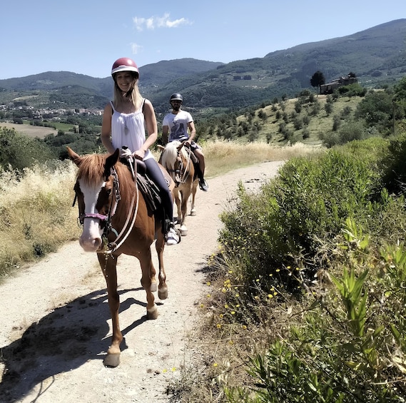 Two people horseback riding on a dirt path surrounded by greenery and hills. 4