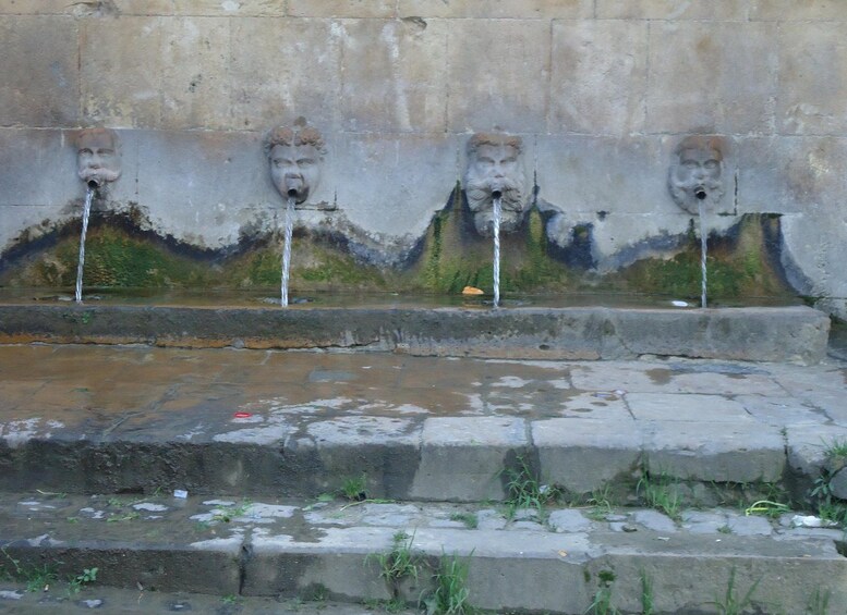 A stone wall with four water spouts, each with a carved face, and a small pool in front. 3