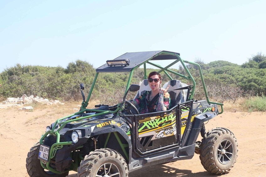 A person in a green and black off-road vehicle with a license plate, parked on a dirt road with sparse vegetation in the background. 1