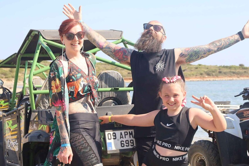 A group of people posing near a green vehicle with a license plate, a body of water, and a hilly landscape in the background. 2