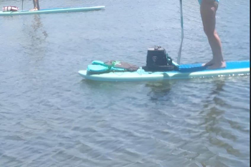 A person standing on a paddleboard in calm water, with a paddle and a bag on the board. 4