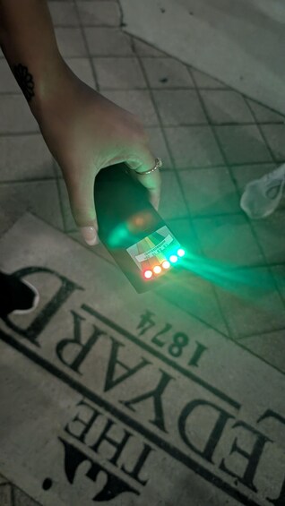 A hand holding a device with a green light, a tattoo on the wrist, and a newspaper with partially visible text. 4