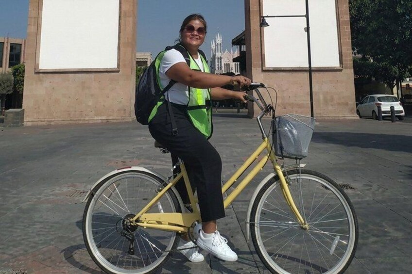 A person riding a yellow bicycle with a basket on the front, wearing a white shirt, black pants, and white sneakers, with a backpack. 2