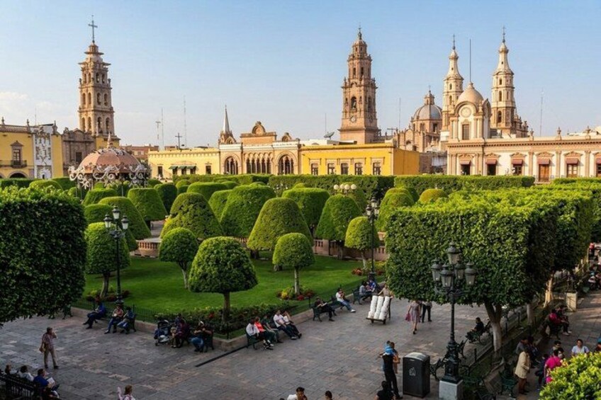 A plaza with trimmed hedges, people sitting and walking, and historic buildings with towers in the background. 4
