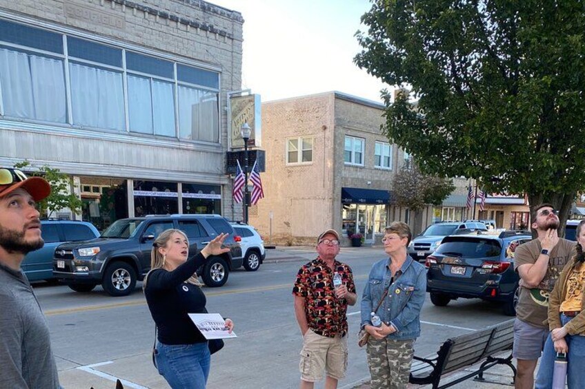 A group of people standing on a sidewalk in an urban area with buildings and parked cars. 1