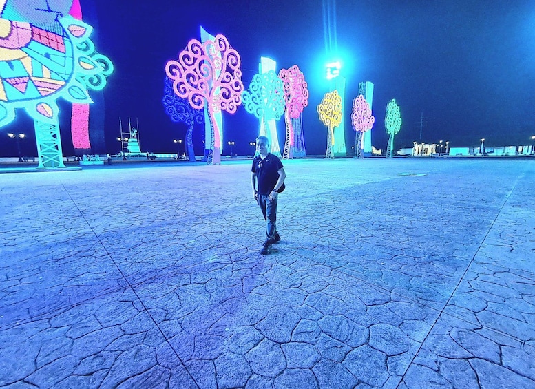 A person standing in a plaza with illuminated trees and a geometric sculpture. 2