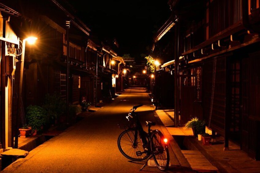 A narrow street at night with buildings on either side, a bicycle parked on the right, and a person walking in the distance. 3