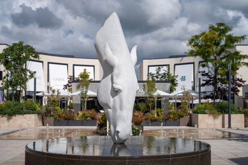 A modern sculpture of a horse's head in a plaza with a fountain, surrounded by buildings and greenery. 1