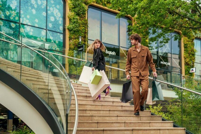 Deux personnes descendant un escalier dans un centre commercial, avec des rampes en verre et des plantes vertes. 5