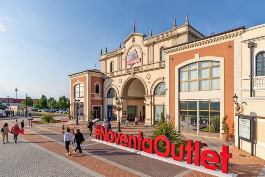 A shopping center entrance with a sign reading 'Noventa Outlet' and a clear sky above. 3