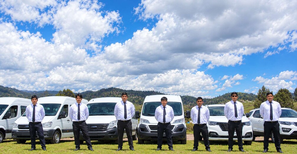 A group of men in business attire standing in front of white vans in a grassy field with a mountainous backdrop. 4
