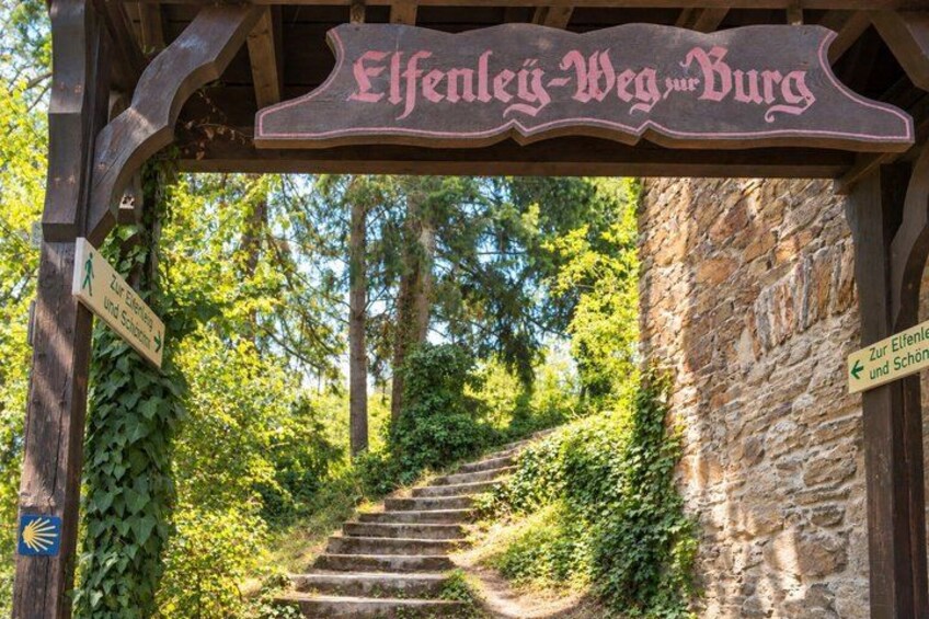 A wooden sign with 'Elfenweg' written on it, a stone wall, and a staircase leading up a hill. 3