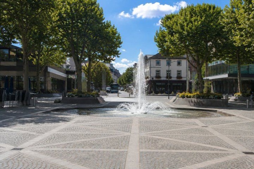 A fountain in a plaza with patterned paving, surrounded by trees and buildings. 3