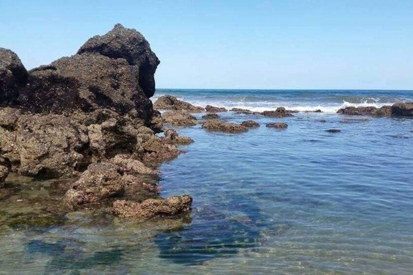 A rocky shoreline with clear water, a large rock formation, and the ocean in the background. 3