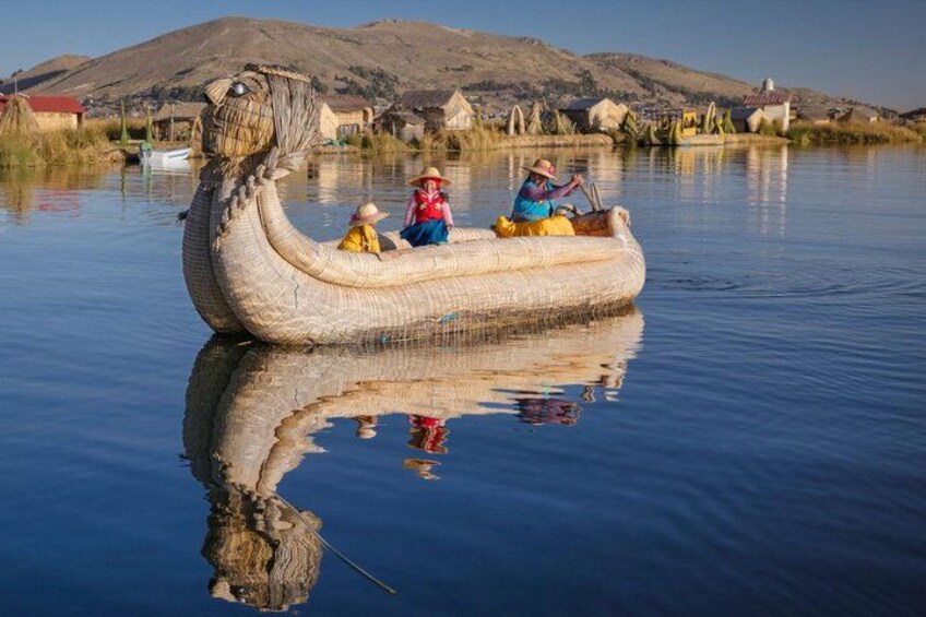A traditional boat with two people, a calm body of water, and houses in the background. 3