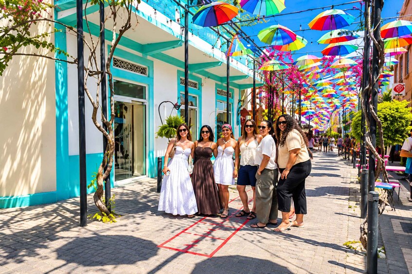 A group of people standing on a street lined with colorful umbrellas and blue buildings. 1